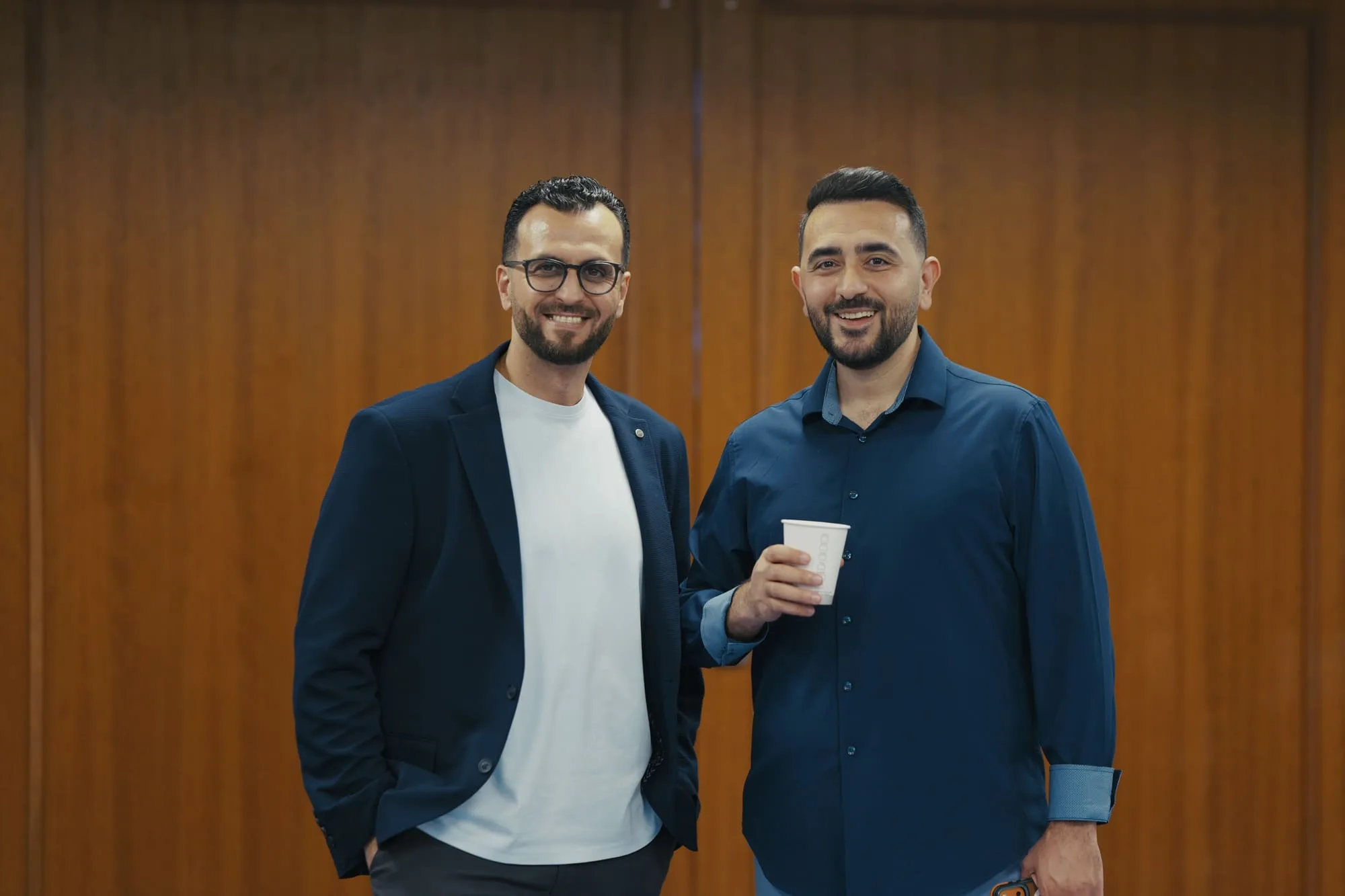 Two men smiling together in front of a warm wood-paneled wall