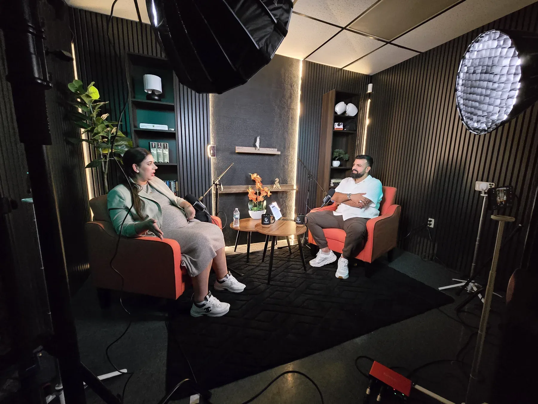 Wide view of the podcast set with two seated speakers, microphones, softboxes, and phones on compact stands